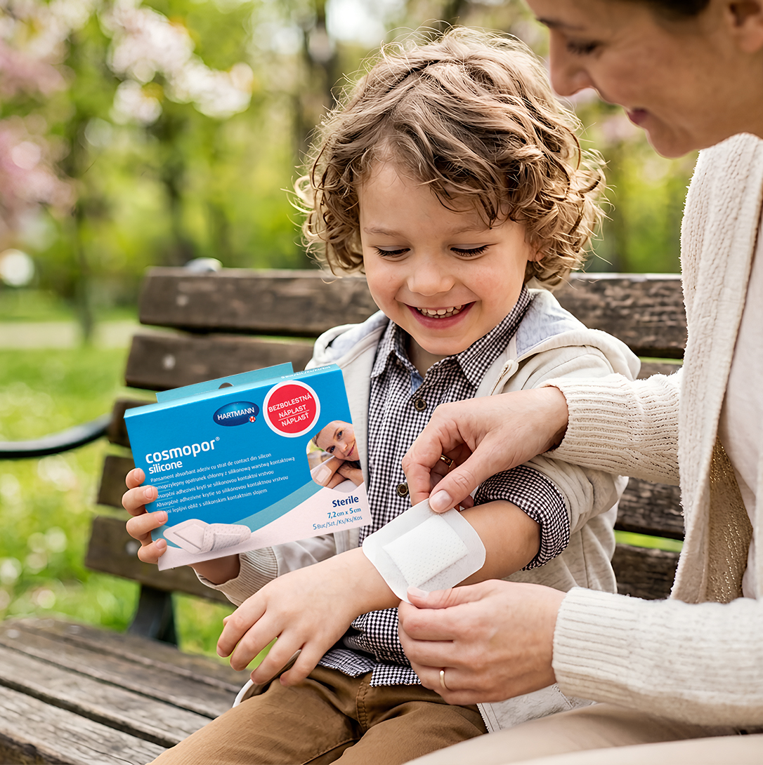 Maminka pomáhá zraněnému dítěti nalepit silikonovou náplast Cosmopor® Silicone