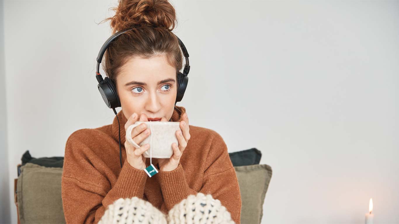 Žena sedí v poklidu na gauči a při poslechu hudby popíjí čaj